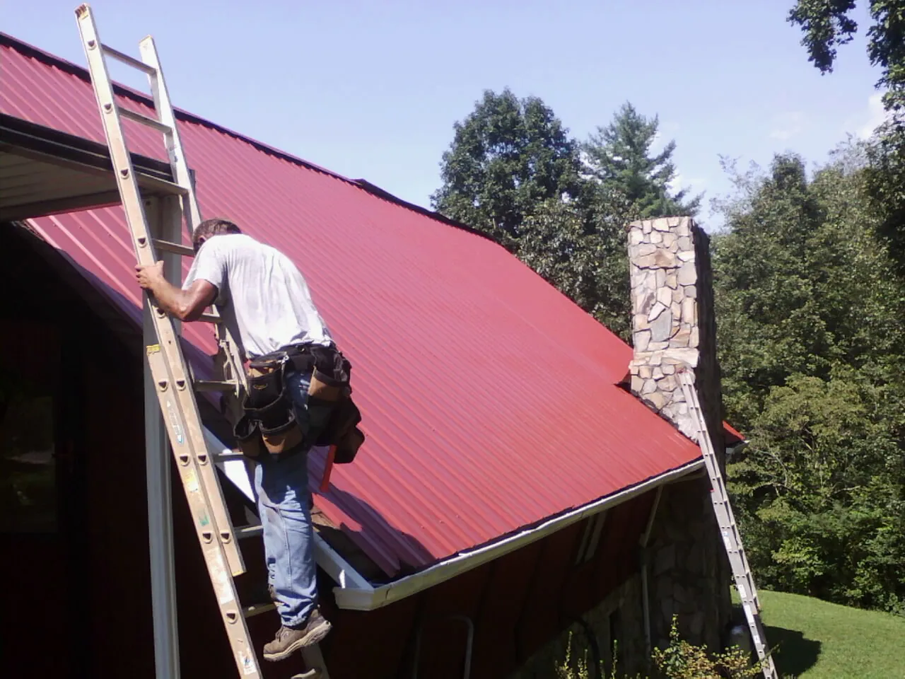 Roofer on a ladder beside a red metal roof and stone chimney — Soffit & Fascia Repair by Roofing Brothers in Bluefield